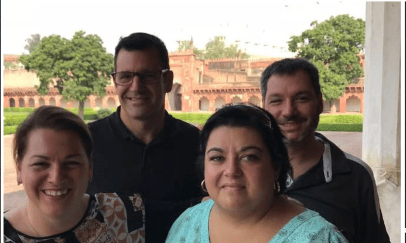 Four travellers smiling together before the red-sandstone walls and green gardens of a Mughal fort in India.