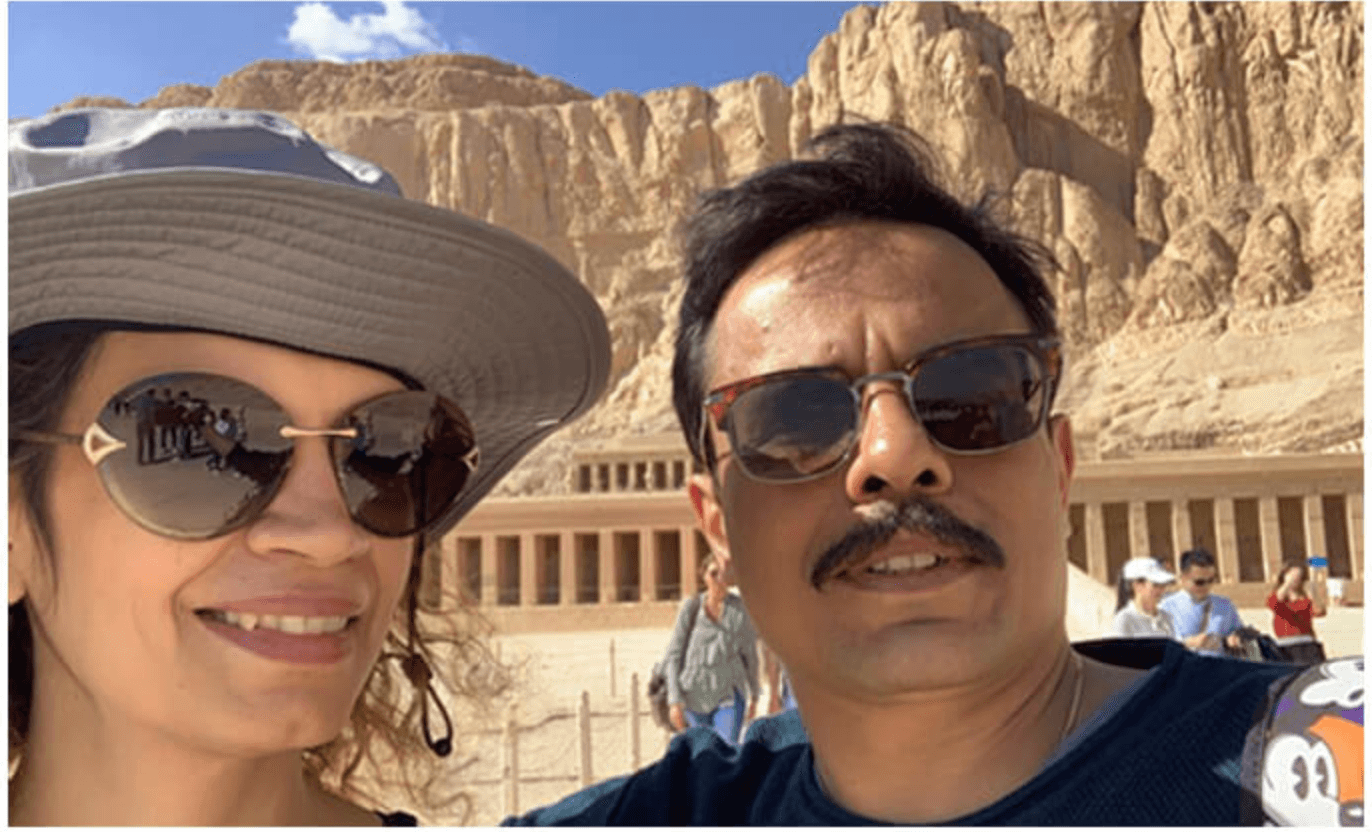 A couple in sun hats and sunglasses before the terraced cliffs of an ancient desert temple.